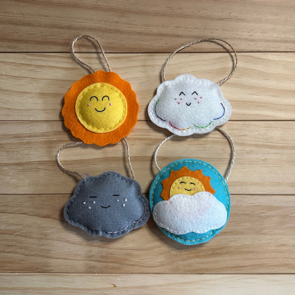 Four small felt cloud, rain cloud, partly sunny and sun ornaments with embroidered faces, hanging from twine loops on a light wood surface.