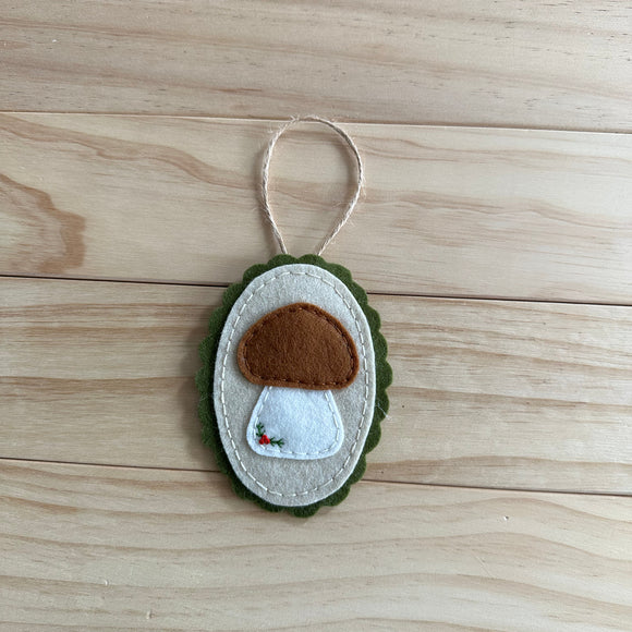 A handmade felt mushroom ornament with a brown cap, white stem, and green scalloped border, hanging from a twine loop on a light wood plank background.