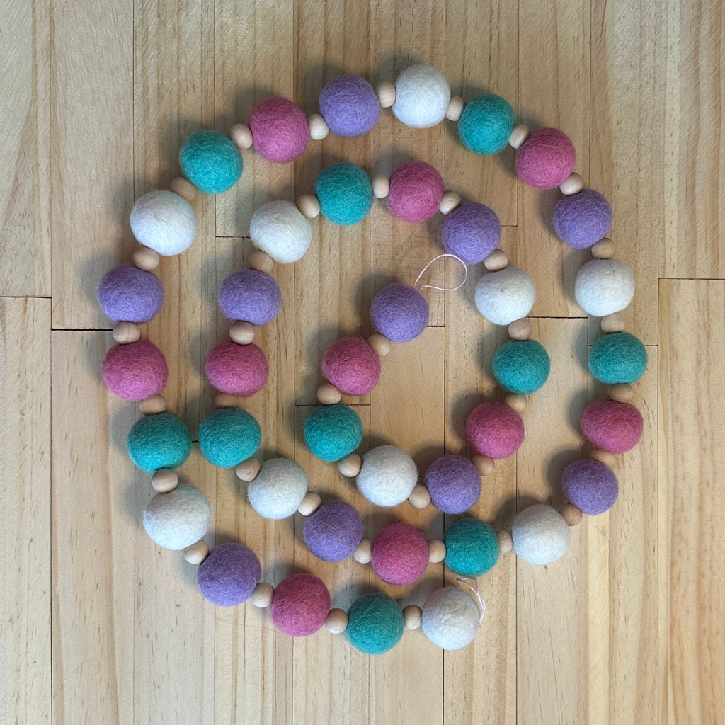A beaded garland made of alternating pastel felt balls in pink, purple, teal, and white, separated by small natural wood beads, arranged in a spiral on a light wood floor.