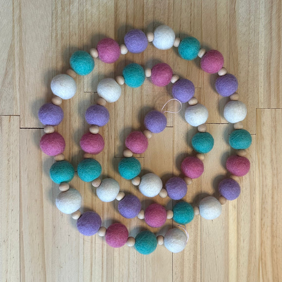 A beaded garland made of alternating pastel felt balls in pink, purple, teal, and white, separated by small natural wood beads, arranged in a spiral on a light wood floor.