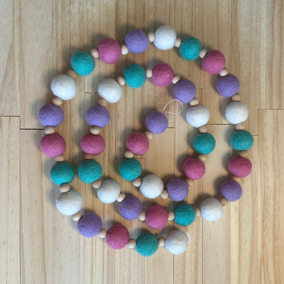 A beaded garland made of alternating pastel felt balls in pink, purple, teal, and white, separated by small natural wood beads, arranged in a spiral on a light wood floor.