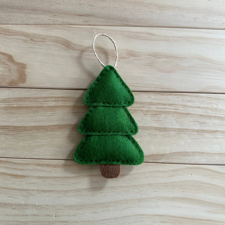 A small felt Christmas tree ornament with green layered foliage and a brown trunk, hanging from a white string on a light wood plank background.