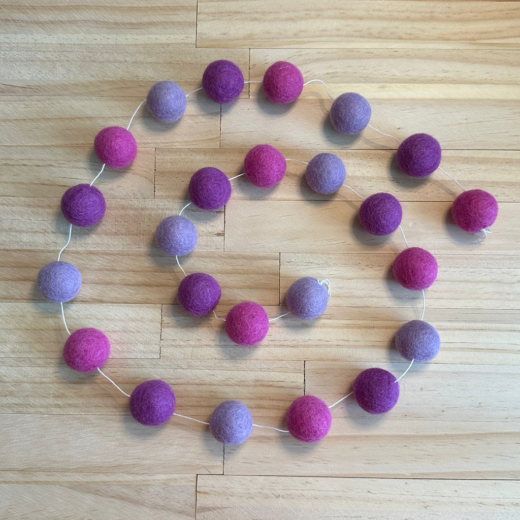 A felt garland made of wool balls in alternating shades of purple and magenta pink, strung along a thin white cord and laid out on a light wood floor.