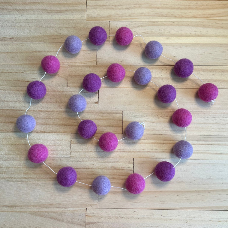 A felt garland made of wool balls in alternating shades of purple and magenta pink, strung along a thin white cord and laid out on a light wood floor.