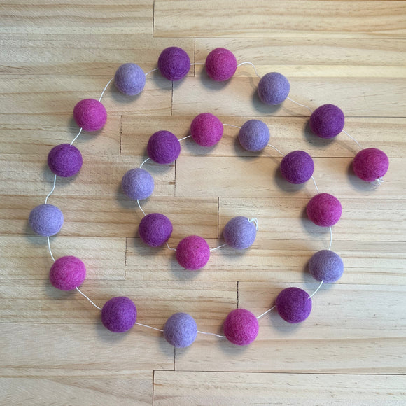 A felt garland made of wool balls in alternating shades of purple and magenta pink, strung along a thin white cord and laid out on a light wood floor.