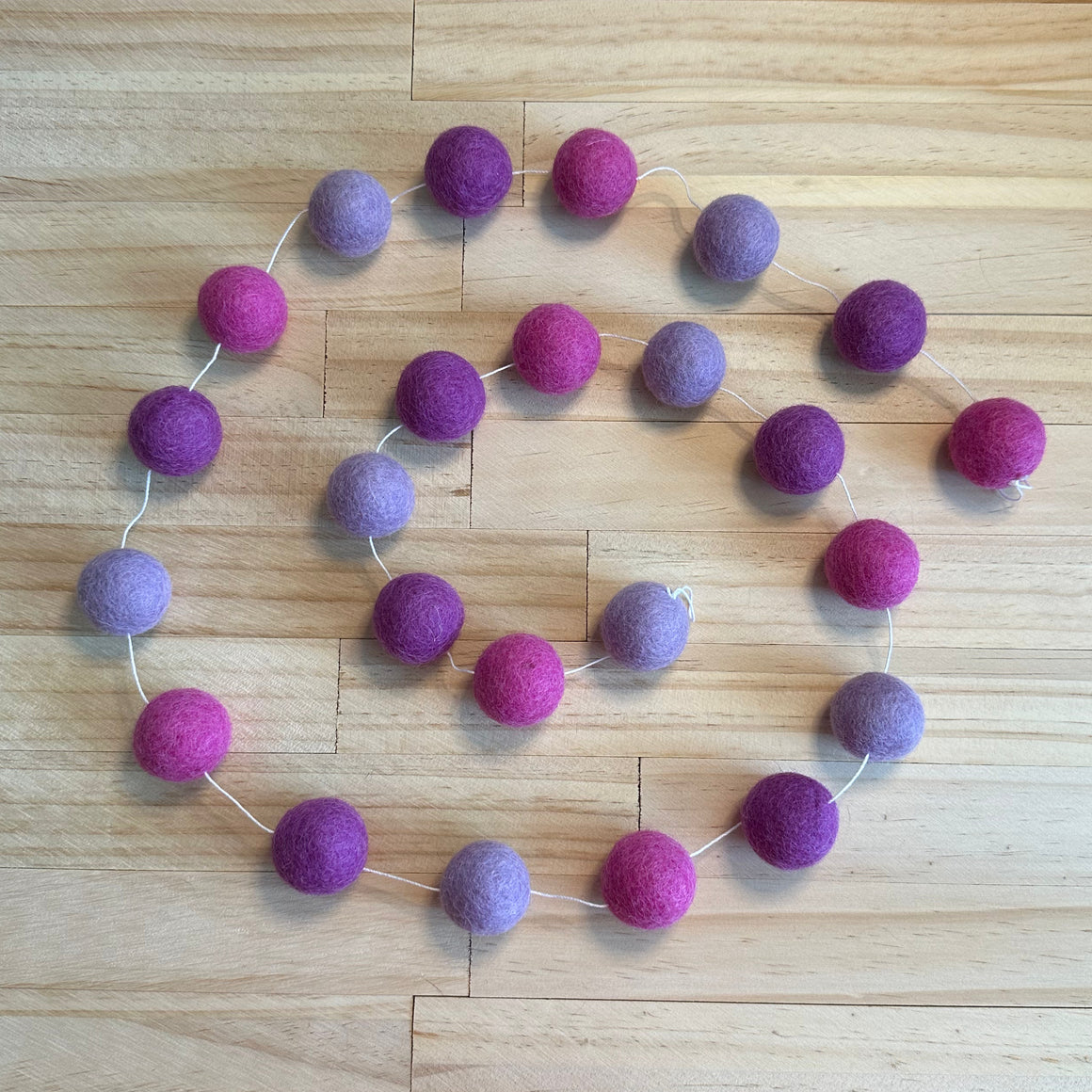 A felt garland made of wool balls in alternating shades of purple and magenta pink, strung along a thin white cord and laid out on a light wood floor.