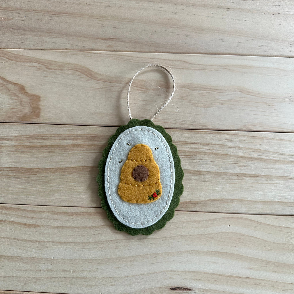 A handmade felt beehive ornament with embroidered bees, and a white hanging loop, resting on a light wood plank surface.