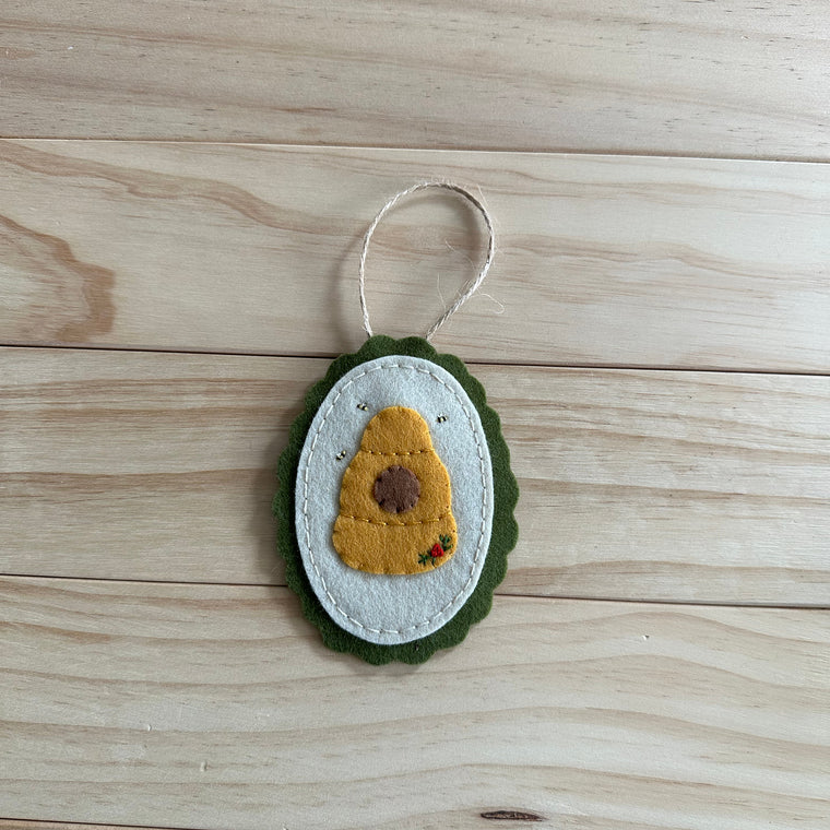 A handmade felt beehive ornament with embroidered bees, and a white hanging loop, resting on a light wood plank surface.