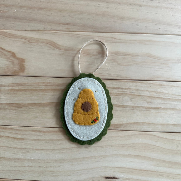 A handmade felt beehive ornament with embroidered bees, and a white hanging loop, resting on a light wood plank surface.