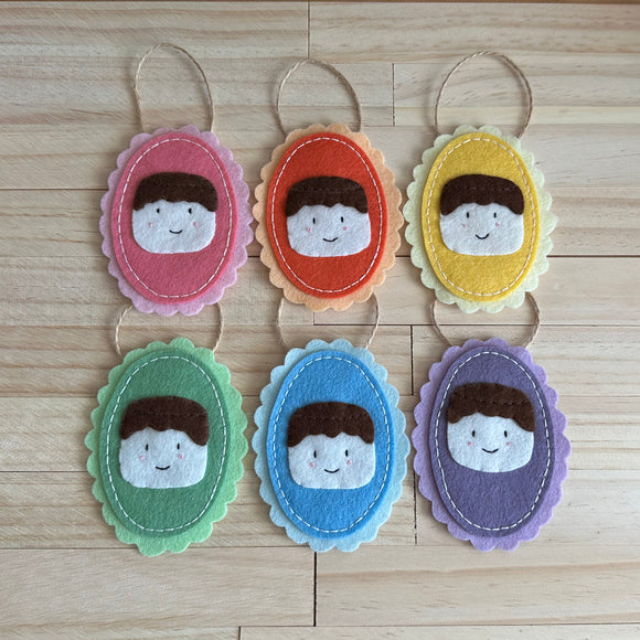 Six handmade oval felt ornaments with scalloped edges, each featuring a chocolate covered marshmallow with a kawaii face and a loop at the top, arranged on a light wood surface. The faces are shown in pink, orange, yellow, green, blue, and purple colorways.